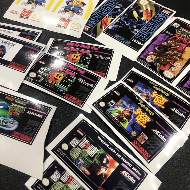 K&ouml;nnte beinhalten: Eine Sammlung von Super Nintendo-Spielekartuschen-Etiketten. Die Etiketten zeigen verschiedene Figuren und Logos, darunter Ms. Pac-Man, Teenage Mutant Ninja Turtles, Scooby-Doo und Frank Thomas Big Hurt Baseball. Die Etiketten sind auf wei&szlig;em Papier mit schwarzem Text und Grafiken gedruckt.