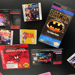 K&ouml;nnte beinhalten: Eine Sammlung von Videospiel-Kartuschen-Etiketten, darunter Contra III: The Alien Wars, The Legend of Zelda: Link's Awakening DX, Maximum Carnage, Castlevania Dracula X, Batman: The Video Game und Street Fighter II. Die Etiketten befinden sich auf einem schwarzen Hintergrund.