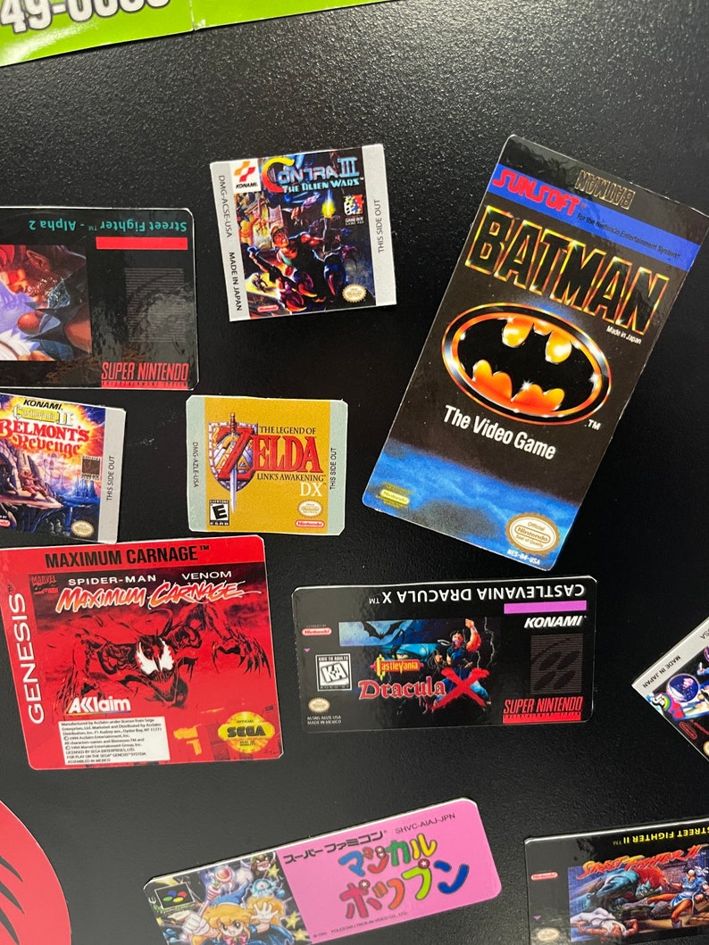K&ouml;nnte beinhalten: Eine Sammlung von Videospiel-Kartuschen-Etiketten, darunter Contra III: The Alien Wars, The Legend of Zelda: Link's Awakening DX, Maximum Carnage, Castlevania Dracula X, Batman: The Video Game und Street Fighter II. Die Etiketten befinden sich auf einem schwarzen Hintergrund.