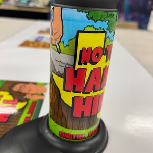 May include: A black and green cylindrical container with a label that says "No Till Hand Hi" and a cartoon image of a hand holding a cigarette. The container is sitting on a black base.