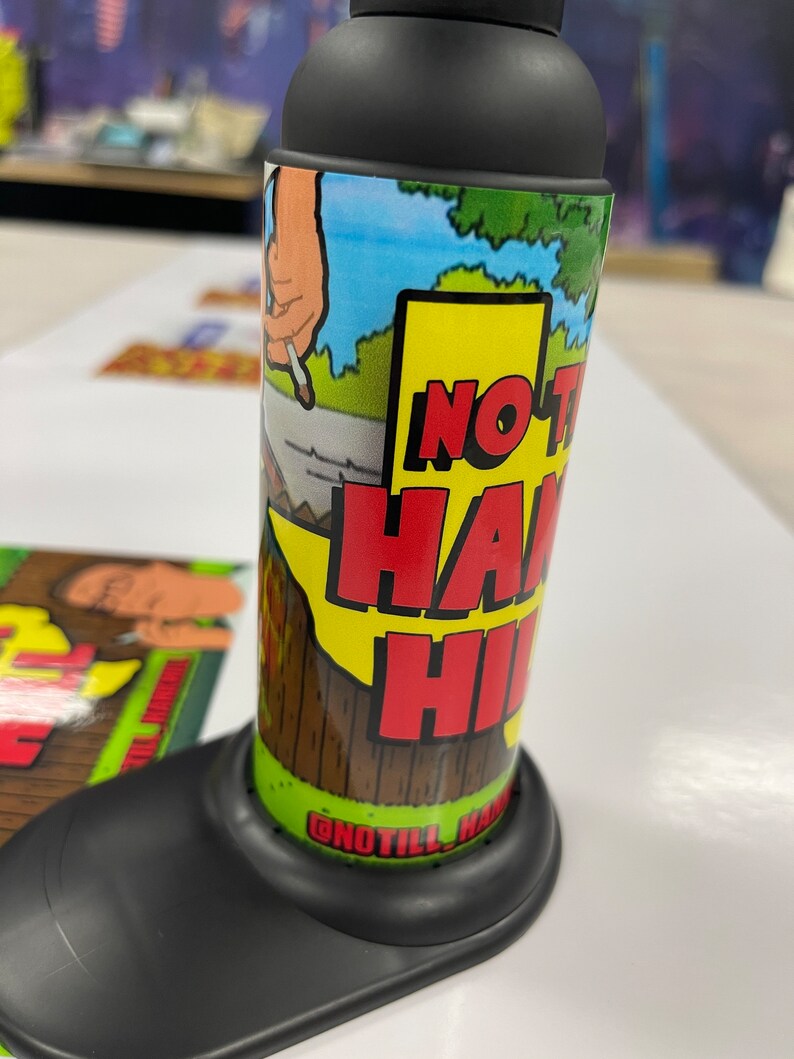 May include: A black and green cylindrical container with a label that says "No Till Hand Hi" and a cartoon image of a hand holding a cigarette. The container is sitting on a black base.