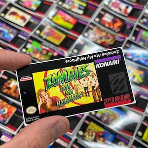 K&ouml;nnte beinhalten: Ein Retro-Videospielmodul f&uuml;r das Super Nintendo Entertainment System. Das Etikett zeigt den Titel "Zombies Ate My Neighbors" mit einer farbenfrohen Grafik von Zombies und erschrockenen Menschen. Das Modul wird in einer Hand gehalten.