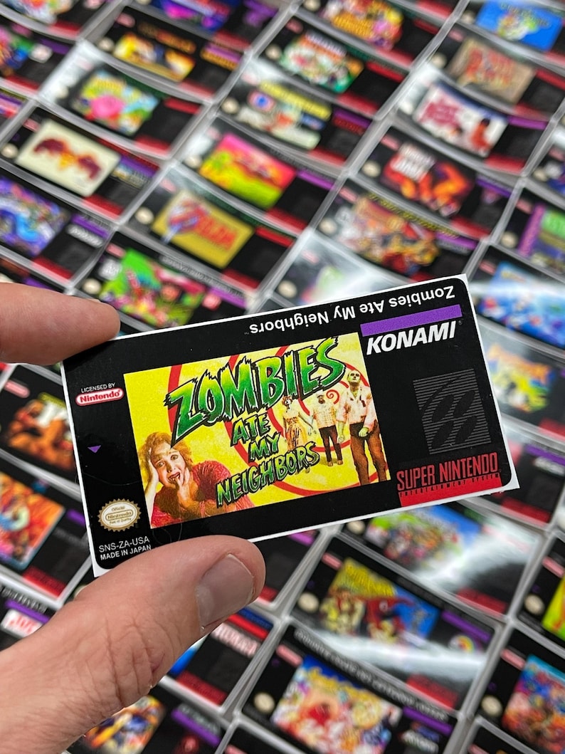 K&ouml;nnte beinhalten: Ein Retro-Videospielmodul f&uuml;r das Super Nintendo Entertainment System. Das Etikett zeigt den Titel "Zombies Ate My Neighbors" mit einer farbenfrohen Grafik von Zombies und erschrockenen Menschen. Das Modul wird in einer Hand gehalten.