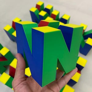Può includere: Un grande logo 'N' stampato in 3D, colorato, con forme geometriche blu, verdi e gialle. Il logo è tenuto in una mano, con altre forme simili sparse sullo sfondo. Il design ricorda una classica console per videogiochi.