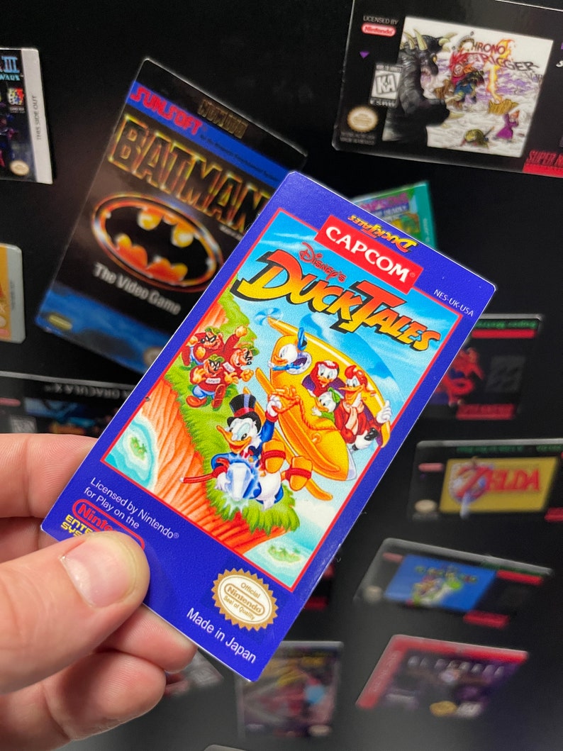K&ouml;nnte beinhalten: Eine blaue und gelbe Nintendo Entertainment System (NES) Spielkassette f&uuml;r DuckTales, mit einem Comicbild von Dagobert Duck und seinen Neffen Tick, Trick und Track auf der Vorderseite. Das Spiel ist von Nintendo lizenziert und in Japan hergestellt.