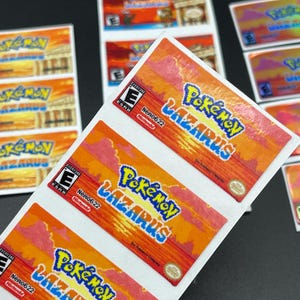 May include: Sticker sheets featuring the "Pokémon Lazarus" game title. The stickers have a vibrant orange, red, and yellow sunset-themed background. The game title is in blue and yellow. The ESRB rating and Nintendo logo are also visible.