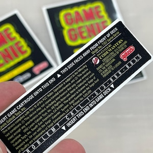 Könnte beinhalten: Nahaufnahme von drei Game Genie Spielkassettenetiketten. Die Etiketten sind schwarz mit rotem und grünem Text. Ein Etikett ist fokussiert und zeigt den Text "INSERT GAME CARTRIDGE ONTO THIS END" und andere Produktinformationen.