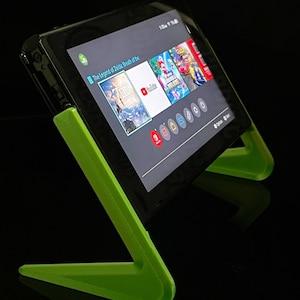 May include: A black Nintendo Switch console is propped up on a bright green stand. The screen displays the game "The Legend of Zelda: Breath of the Wild" and other game icons. The stand is angled for optimal viewing.