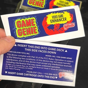 Könnte beinhalten: Ein Game Genie Video-Spiel-Enhancer mit blauem Hintergrund und den Worten "GAME GENIE" in Grün und Gelb. Der Text "VIDEO GAME ENHANCER" ist in Gelb mit einer roten, muskulösen Figur. Die Rückseite der Karte hat Text und das Galoob-Logo.