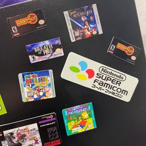 K&ouml;nnte beinhalten: Eine Sammlung von Videospiel-Kartuschen-Etiketten, darunter Super Mario Bros. Deluxe, Golden Sun, Breath of Fire, Super Metroid, Chrono Trigger und Die Simpsons: Flucht aus dem Todeslager. Die Etiketten befinden sich auf einer schwarzen Oberfl&auml;che mit einem Nintendo Super Famicom-Logo.