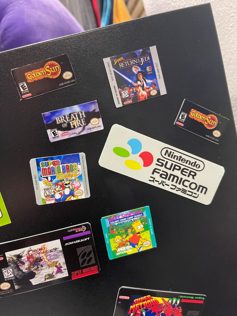 K&ouml;nnte beinhalten: Eine Sammlung von Videospiel-Kartuschen-Etiketten, darunter Super Mario Bros. Deluxe, Golden Sun, Breath of Fire, Super Metroid, Chrono Trigger und Die Simpsons: Flucht aus dem Todeslager. Die Etiketten befinden sich auf einer schwarzen Oberfl&auml;che mit einem Nintendo Super Famicom-Logo.