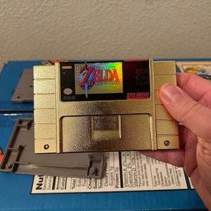 K&ouml;nnte beinhalten: Eine goldfarbene Super Nintendo-Spielkassette f&uuml;r "The Legend of Zelda: A Link to the Past". Die Kassette hat eine goldene Oberfl&auml;che und zeigt das Logo und den Titel des Spiels. Die Kassette wird in einer Hand gehalten.