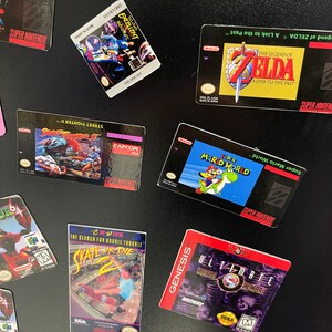 K&ouml;nnte beinhalten: Eine Sammlung von Vintage-Videospielkassetten f&uuml;r die Konsolen Super Nintendo, Nintendo 64 und Sega Genesis. Die Kassetten sind auf einer schwarzen Oberfl&auml;che angeordnet. Die Kassetten beinhalten Titel wie "The Legend of Zelda: A Link to the Past", "Super Mario World", "Street Fighter II", "Castlevania", "Skate or Die 2" und "Ultimate Mortal Kombat 3".