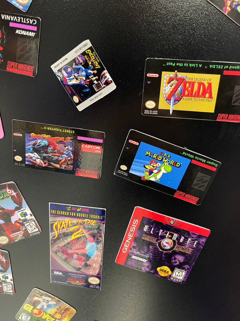 K&ouml;nnte beinhalten: Eine Sammlung von Vintage-Videospielkassetten f&uuml;r die Konsolen Super Nintendo, Nintendo 64 und Sega Genesis. Die Kassetten sind auf einer schwarzen Oberfl&auml;che angeordnet. Die Kassetten beinhalten Titel wie "The Legend of Zelda: A Link to the Past", "Super Mario World", "Street Fighter II", "Castlevania", "Skate or Die 2" und "Ultimate Mortal Kombat 3".