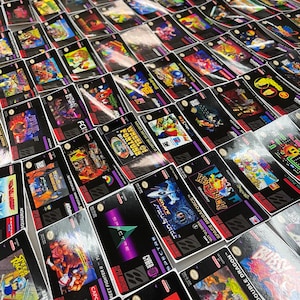 K&ouml;nnte beinhalten: Eine Sammlung von Super Nintendo-Spielkassetten mit verschiedenen Spieletiteln und farbenfrohen Cover-Artworks. Die Kassetten sind eng angeordnet und zeigen eine Vielzahl von Spielgenres und -designs. Das Bild wurde aus einem hohen Winkel aufgenommen.