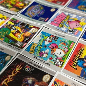K&ouml;nnte beinhalten: Eine Sammlung von bunten Game Boy-Spielekartuschen-Etiketten. Die Etiketten zeigen verschiedene Charaktere und Titel, darunter Kirby's Dream Land 2, Mega Man und Primal Rage. Die Etiketten sind auf silbernem Hintergrund mit schwarzem Text gedruckt.