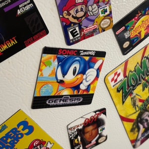K&ouml;nnte beinhalten: Eine Sammlung von Vintage-Videospiel-Kassettenmagneten mit Charakteren wie Mario, Sonic the Hedgehog und James Bond. Die Magnete sind bunt und zeigen die Spieltitel "Super Smash Bros", "Sonic the Hedgehog", "Super Mario 3", "GoldenEye 007", "Mortal Kombat", "Earthbound" und "Zombie Ate My Neighbors".
