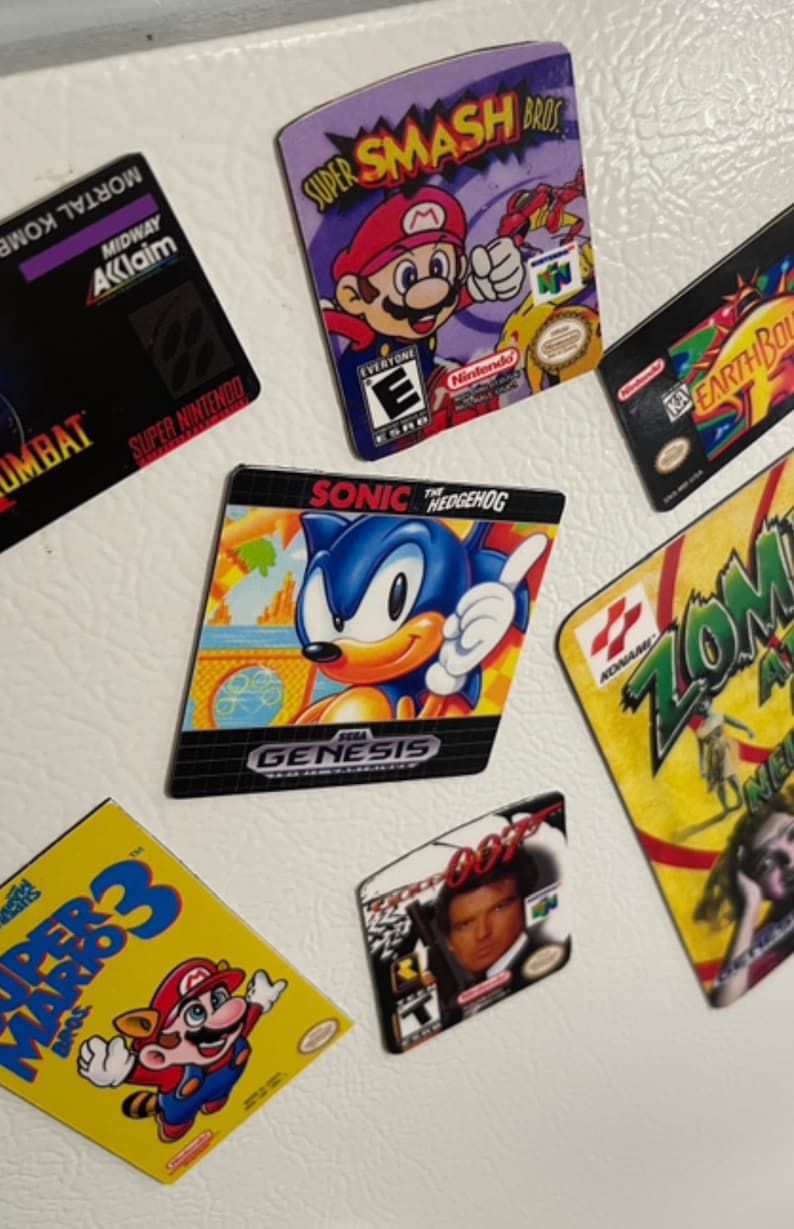 K&ouml;nnte beinhalten: Eine Sammlung von Vintage-Videospiel-Kassettenmagneten mit Charakteren wie Mario, Sonic the Hedgehog und James Bond. Die Magnete sind bunt und zeigen die Spieltitel "Super Smash Bros", "Sonic the Hedgehog", "Super Mario 3", "GoldenEye 007", "Mortal Kombat", "Earthbound" und "Zombie Ate My Neighbors".