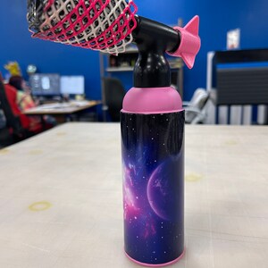May include: A pink and black hair styling tool with a nozzle and a removable, pink and white, mesh attachment. The tool has a galaxy-themed design with a pink and blue nebula and a planet.