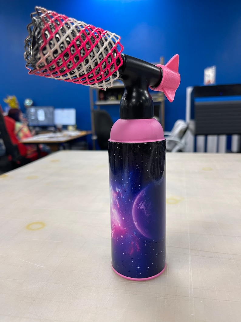 May include: A pink and black hair styling tool with a nozzle and a removable, pink and white, mesh attachment. The tool has a galaxy-themed design with a pink and blue nebula and a planet.