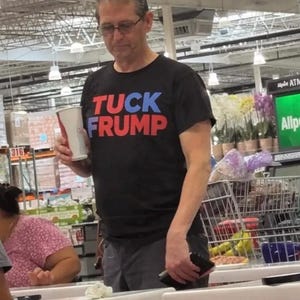 May include: A black t-shirt with the words "TUCK FRUMP" in blue and red block letters. The person in the image is wearing glasses and holding a cup. The background shows a store setting.