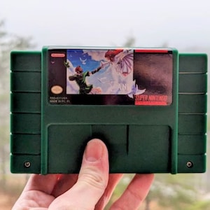 K&ouml;nnte beinhalten: Eine gr&uuml;ne Super Nintendo-Spielkassette mit einem farbenfrohen Etikett, das eine Figur in gr&uuml;ner Kleidung und eine Frau in einem wei&szlig;en Kleid zeigt. Das Spiel tr&auml;gt den Titel "DANI". Die Kassette wird von einer Hand gehalten.