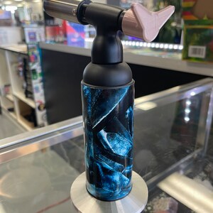 May include: A black and blue torch lighter with a pink tip and a silver nozzle. The lighter is on a silver stand.