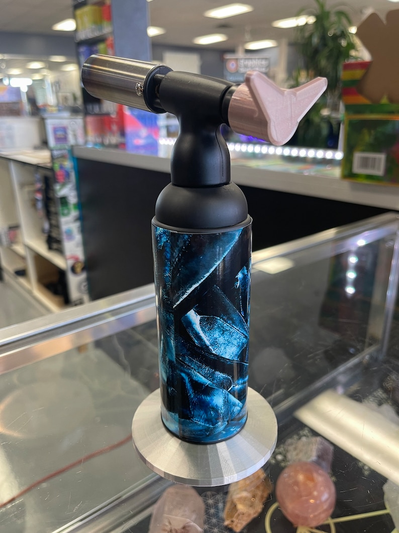 May include: A black and blue torch lighter with a pink tip and a silver nozzle. The lighter is on a silver stand.