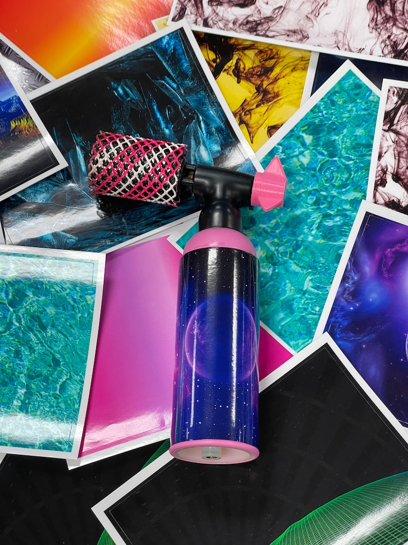 May include: A pink and black torch with a black nozzle and a pink handle. The torch is decorated with a galaxy design with a large purple planet and stars. The torch is surrounded by colorful patterned paper.