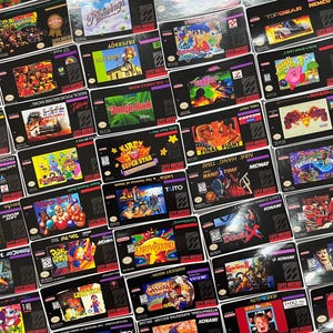 K&ouml;nnte beinhalten: Eine Sammlung von Super Nintendo-Spielkassetten, jede mit einzigartigem Cover-Art. Titel sind u.a. "Donkey Kong Country", "Pilotwings", "Paperboy", "Jungle Book", "Kirby Super Star", "Final Fight", "Earthbound" und "Harvest Moon". Die Kassetten sind schwarz mit farbenfrohen Spielgrafiken.
