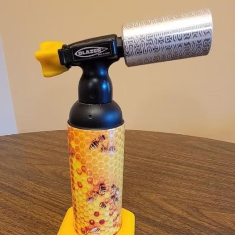 May include: A black and yellow torch with a honeycomb pattern and bees. The torch has a silver nozzle with a textured pattern. The brand name "BLAZER" is printed on the side of the torch.