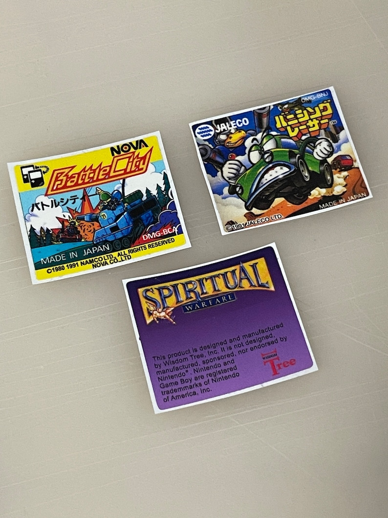 K&ouml;nnte beinhalten: Drei Spielkartuschen-Etiketten f&uuml;r den Nintendo Game Boy. Das erste Etikett ist gelb und blau mit dem Text "NOVA Battle City" und "バトルシティ" in Japanisch. Das zweite Etikett ist gr&uuml;n und blau mit dem Text "JALECO バニシングレーサー" in Japanisch. Das dritte Etikett ist lila mit dem Text "SPIRITUAL WARFARE" und "Dieses Produkt wurde von Wisdom Tree, Inc. entworfen und hergestellt. Es wurde weder von Nintendo entworfen, hergestellt, gesponsert noch unterst&uuml;tzt. Nintendo und Game Boy sind eingetragene Marken von Nintendo of America, Inc."