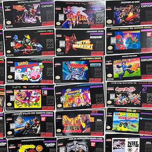K&ouml;nnte beinhalten: Eine Sammlung von Super Nintendo-Spielkassetten mit verschiedenen Titeln und farbenfrohen Box-Art. Zu den Spielen geh&ouml;ren "Kirby's Dream Course", "Batman Returns" und "Mighty Morphin Power Rangers". Die Kassetten sind auf einer schwarzen Oberfl&auml;che angeordnet.