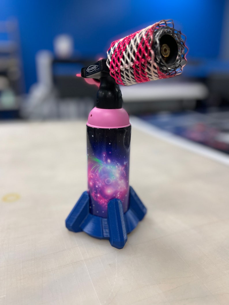 May include: A pink and black torch with a galaxy print design and a blue base. The torch has a white and red mesh covering the nozzle.