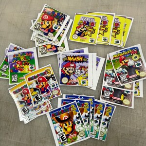 K&ouml;nnte beinhalten: Eine Sammlung von Nintendo 64-Spielekartuschen-Etiketten mit verschiedenen Mario-Spielen, darunter Mario Party, Super Smash Bros, Mario Kart und Paper Mario. Die Etiketten sind auf wei&szlig;em Papier mit bunten Grafiken und Text gedruckt.