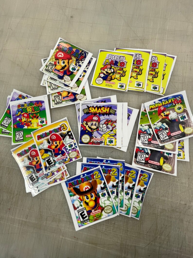 K&ouml;nnte beinhalten: Eine Sammlung von Nintendo 64-Spielekartuschen-Etiketten mit verschiedenen Mario-Spielen, darunter Mario Party, Super Smash Bros, Mario Kart und Paper Mario. Die Etiketten sind auf wei&szlig;em Papier mit bunten Grafiken und Text gedruckt.