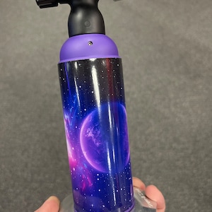 May include: A purple and black torch with a space-themed design. The torch has a silver nozzle and a black handle. The design features a galaxy with stars and a planet.