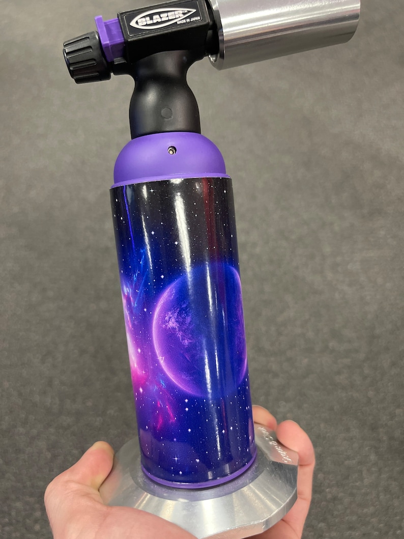 May include: A purple and black torch with a space-themed design. The torch has a silver nozzle and a black handle. The design features a galaxy with stars and a planet.