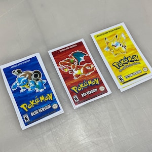 May include: Three Pokémon game cartridges: Blue Version, Red Version, and Yellow Version. The Blue Version features a blue Blastoise, the Red Version shows Charizard, and the Yellow Version has Pikachu. Each cartridge has the Pokémon logo and version name.