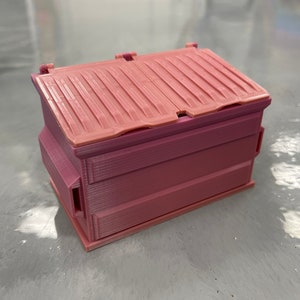 May include: A small, pink, rectangular container with a hinged lid. The container has a ridged top and appears to be 3D-printed. The color is a gradient of pink hues, with a darker shade at the base and a lighter shade on the lid.