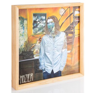 Op de afbeelding: Een ingelijste albumhoes met een man met lang bruin haar, die een lichtblauw overhemd met knopen en een blauwe spijkerbroek draagt. De achtergrond is een gele kamer met een raam en een trap. De albumhoes is getiteld "HOZIER".