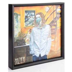 Op de afbeelding: Een ingelijste albumhoes met een man met lang haar die in een kamer staat met een gele muur, een raam en een trap. De albumhoes is getiteld "Hozier".