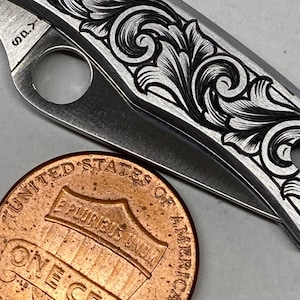 Puede incluir: Un peque&ntilde;o cuchillo plegable plateado con un patr&oacute;n floral grabado. Una moneda de un centavo de cobre est&aacute; al lado del cuchillo. La hoja est&aacute; parcialmente abierta. El cuchillo mide aproximadamente 7,6 cm de largo. El cuchillo tiene un patr&oacute;n floral grabado.