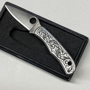 Puede incluir: Una peque&ntilde;a navaja plateada con un patr&oacute;n floral decorativo grabado en el mango. La hoja est&aacute; abierta, revelando un filo afilado. La navaja se presenta en una caja negra, mostrando el dise&ntilde;o y la artesan&iacute;a.
