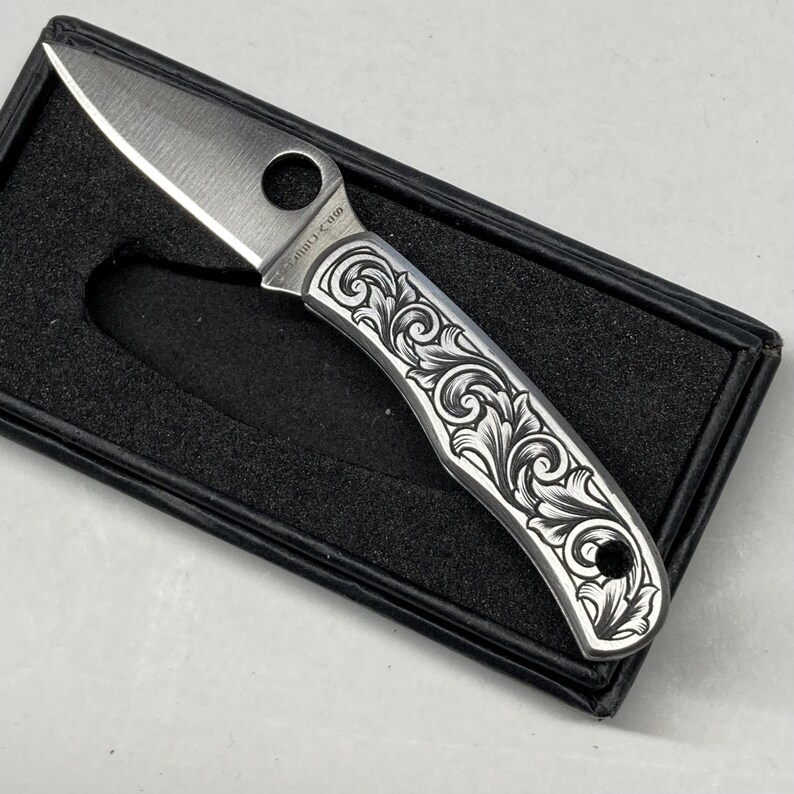 Puede incluir: Una peque&ntilde;a navaja plateada con un patr&oacute;n floral decorativo grabado en el mango. La hoja est&aacute; abierta, revelando un filo afilado. La navaja se presenta en una caja negra, mostrando el dise&ntilde;o y la artesan&iacute;a.