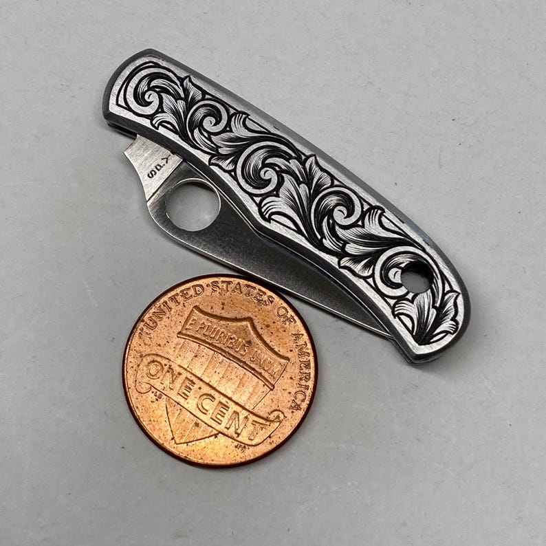 Puede incluir: Un peque&ntilde;o cuchillo plegable plateado con un patr&oacute;n floral grabado. Una moneda de un centavo de cobre est&aacute; al lado del cuchillo. La hoja est&aacute; parcialmente abierta. El cuchillo mide aproximadamente 7,6 cm de largo. El cuchillo tiene un patr&oacute;n floral grabado.