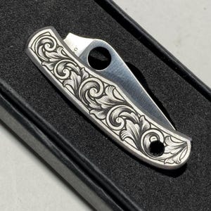 Puede incluir: Una peque&ntilde;a navaja plateada con un patr&oacute;n floral grabado y ornamentado. La hoja est&aacute; parcialmente abierta, revelando un filo de acero pulido. La navaja est&aacute; en una caja negra.