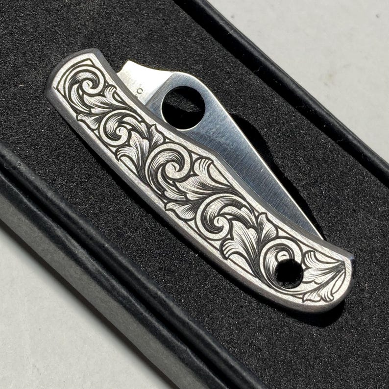 Puede incluir: Una peque&ntilde;a navaja plateada con un patr&oacute;n floral grabado y ornamentado. La hoja est&aacute; parcialmente abierta, revelando un filo de acero pulido. La navaja est&aacute; en una caja negra.