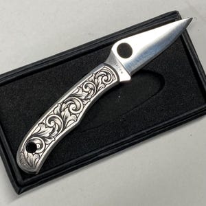 Puede incluir: Una navaja de bolsillo plateada con un dise&ntilde;o floral decorativo grabado en el mango. La hoja es afilada y puntiaguda. La navaja se presenta en una caja negra.