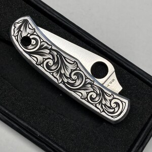 Puede incluir: Una navaja plateada con un intrincado dise&ntilde;o floral negro grabado en el mango. La hoja est&aacute; parcialmente abierta. La navaja se presenta en una caja negra. El dise&ntilde;o es detallado.